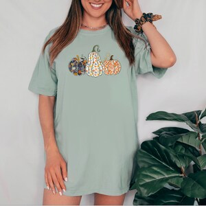 May include: A light green t-shirt with a graphic of four patterned pumpkins. The pumpkins are in a row and are all different colors and patterns.