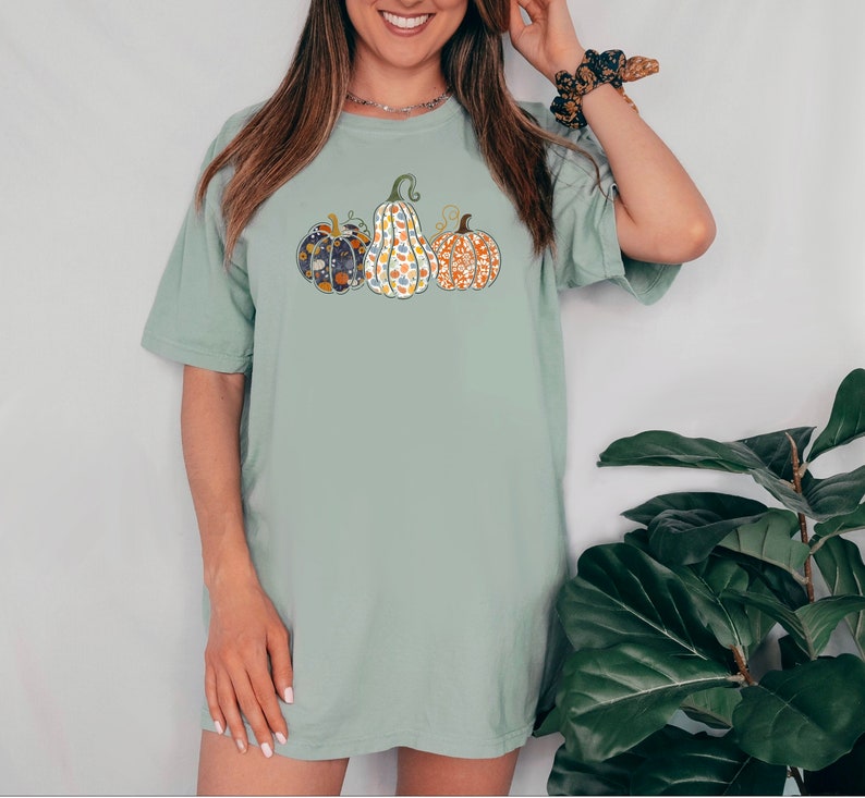 May include: A light green t-shirt with a graphic of four patterned pumpkins. The pumpkins are in a row and are all different colors and patterns.