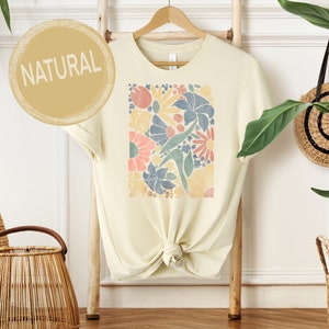 May include: A cream colored t-shirt with a floral print in shades of blue, pink, yellow, and green. The text "NATURAL" is printed on a circle in the top left corner of the image.