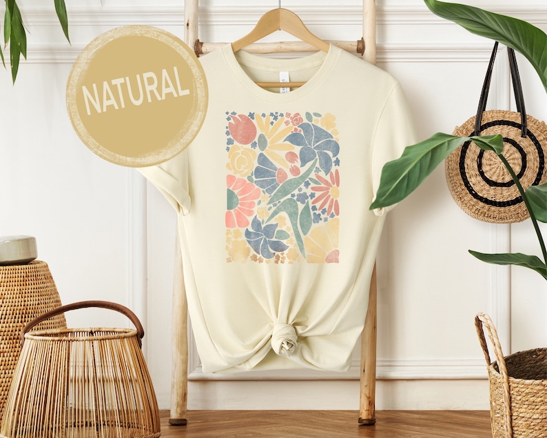 May include: A cream colored t-shirt with a floral print in shades of blue, pink, yellow, and green. The text "NATURAL" is printed on a circle in the top left corner of the image.