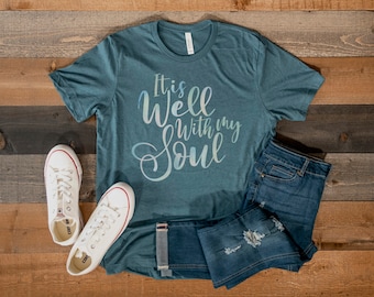 May include: Teal blue t-shirt with the text "It is well with my soul" in a white, blue, and pink gradient. The shirt is laying on a wooden surface with a pair of white canvas shoes and a pair of blue jeans.