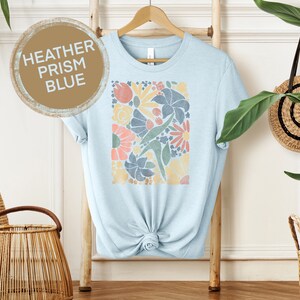 May include: A light blue t-shirt with a floral print in shades of pink, yellow, green, and blue. The print is a square design with a variety of flowers and leaves. The t-shirt is on a hanger and has the words "HEATHER PRISM BLUE" written on a brown circle.