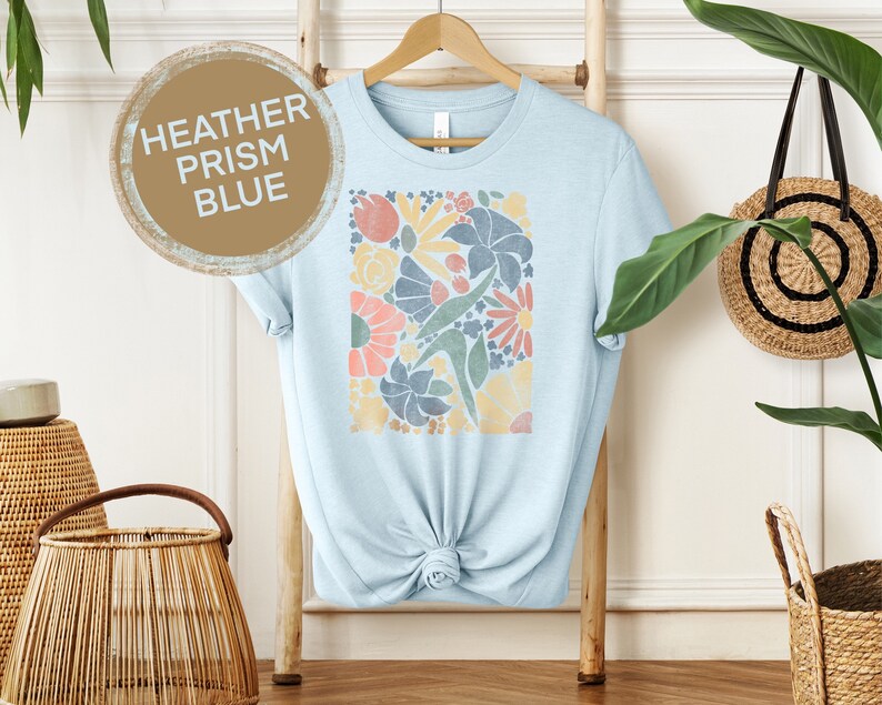 May include: A light blue t-shirt with a floral print in shades of pink, yellow, green, and blue. The print is a square design with a variety of flowers and leaves. The t-shirt is on a hanger and has the words "HEATHER PRISM BLUE" written on a brown circle.