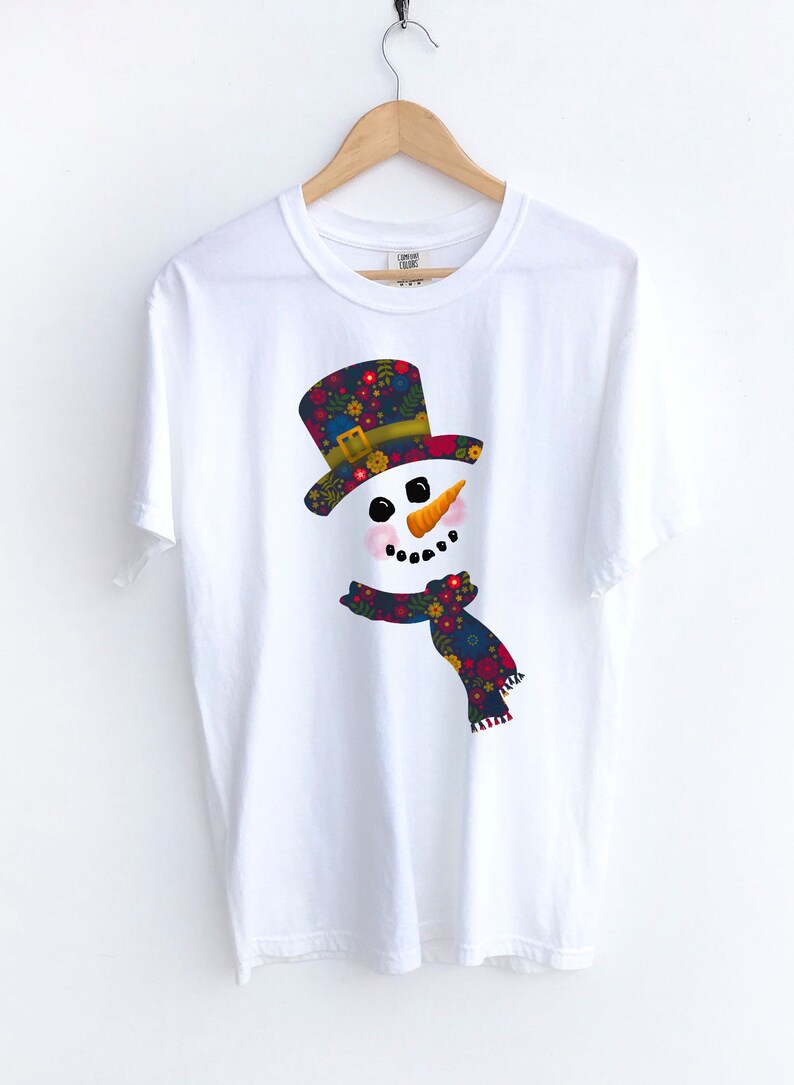 Puede incluir: Camiseta blanca con un gr&aacute;fico de mu&ntilde;eco de nieve colorido. El mu&ntilde;eco de nieve tiene un sombrero y una bufanda con estampado floral.