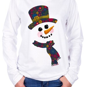 Puede incluir: Camiseta blanca de manga larga con un dise&ntilde;o de mu&ntilde;eco de nieve floral colorido. El mu&ntilde;eco de nieve lleva un sombrero negro con una banda verde y una bufanda floral.
