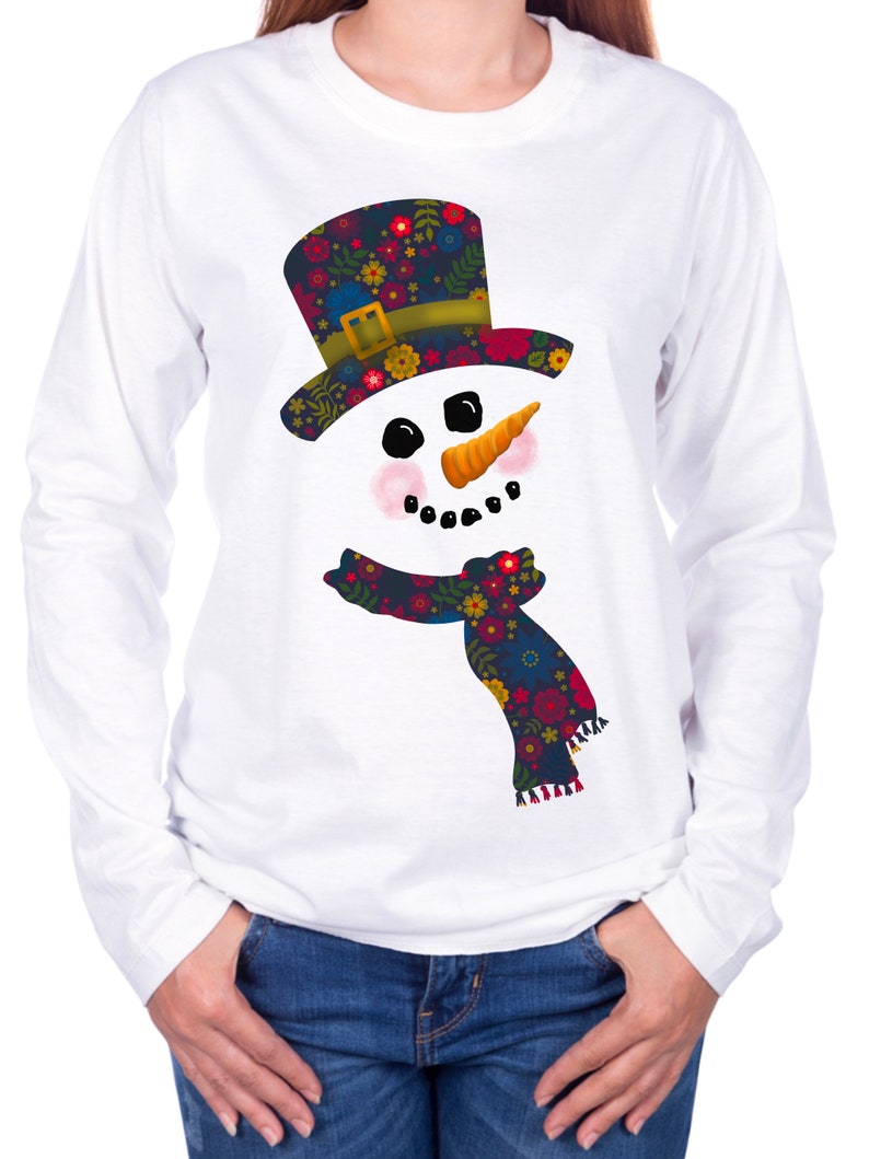 Puede incluir: Camiseta blanca de manga larga con un dise&ntilde;o de mu&ntilde;eco de nieve floral colorido. El mu&ntilde;eco de nieve lleva un sombrero negro con una banda verde y una bufanda floral.