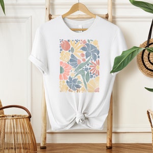 May include: White t-shirt with a colorful floral print design. The print features a variety of flowers and leaves in shades of blue, yellow, pink, and green.
