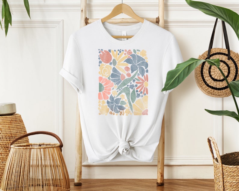 May include: White t-shirt with a colorful floral print design. The print features a variety of flowers and leaves in shades of blue, yellow, pink, and green.