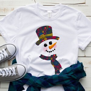 Puede incluir: Camiseta blanca con un dise&ntilde;o de mu&ntilde;eco de nieve. El mu&ntilde;eco de nieve lleva un sombrero y una bufanda con estampado floral.