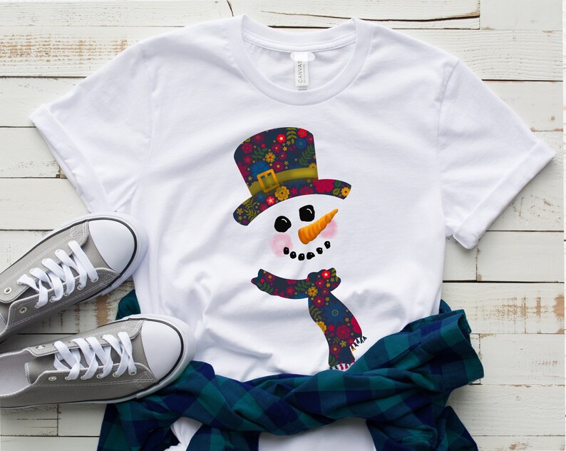 Puede incluir: Camiseta blanca con un dise&ntilde;o de mu&ntilde;eco de nieve. El mu&ntilde;eco de nieve lleva un sombrero y una bufanda con estampado floral.