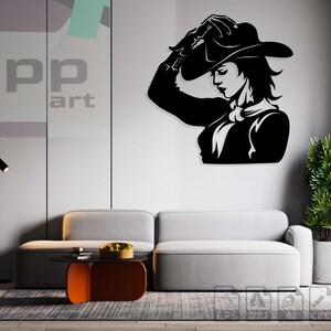 以下が含まれることがあります： カウボーイハットをかぶった女性の白黒のウォールアートで、「yupp art」の文字が入っています。アートワークは、灰色のソファの上にある白い壁に取り付けられています。ミニマリストデザインのモダンなリビングルームの設定。