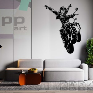 May include: Black and white silhouette of a person riding a motorcycle with arms raised in the air. The image is on a white wall in a living room setting with a gray couch and a black floor lamp.