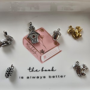 Puede incluir: Un plato de cerámica blanco con un diseño de libro rosa y el texto "the book is always better". El plato contiene varios amuletos plateados y dorados, incluyendo una casita de pájaros, una bellota, una ardilla y una piña.