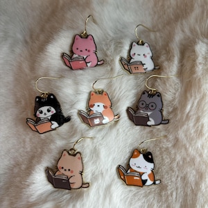 May include: Six pairs of cat-themed earrings. Each earring features a cartoon cat reading a book. The cats are different colors, including pink, white, black, orange, gray, and calico.