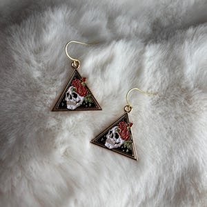 May include: Gold triangle earrings with a black background and a white skull with red roses. The skull is surrounded by a gold outline.