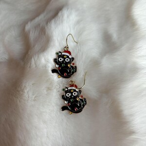 May include: A pair of gold-tone earrings featuring black cat charms wearing Santa hats and holding Christmas lights.