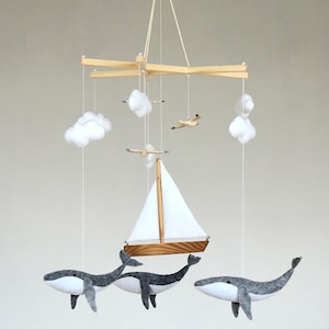 May include: A hanging mobile featuring a wooden sailboat with a white sail, three grey and white felt whales, two small wooden aeroplanes, and several white felt clouds suspended from a wooden cross frame by thin white strings.