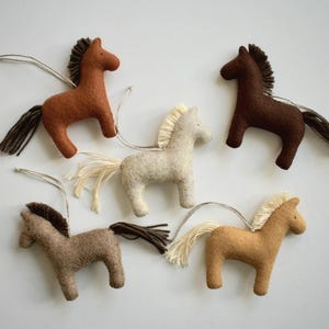 May include: Five felt horse ornaments in various shades of brown, tan, and cream. Each horse has a dark mane and tail, with a string for hanging. The horses are arranged on a white background.