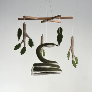 以下が含まれることがあります： 緑のフェルトの蛇、枝、葉がぶら下がっている木製モビール。モビールは白い紐で吊り下げられています。