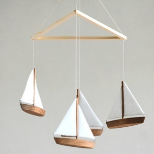 Puede incluir: Un m&oacute;vil de madera con tres veleros blancos colgando de un marco triangular de madera. Los veleros est&aacute;n hechos de madera y tienen velas de tela blanca.