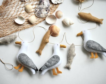 May include: A felt garland with three white seagulls, each holding a felt fish in their beaks. The garland is strung with twine and has a brown and white fishing net in the background.
