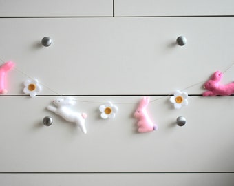 Puede incluir: Una guirnalda con conejos de fieltro rosa y blanco y flores de fieltro blanco con centros amarillos.