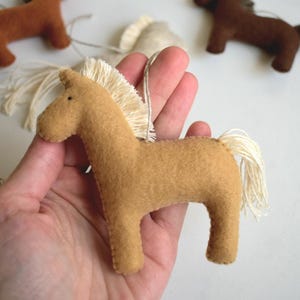 May include: Tan felt horse ornament with a cream-colored mane and tail. The horse is held in a hand, with other similar ornaments in the background. These ornaments are likely for a nursery or children's room.