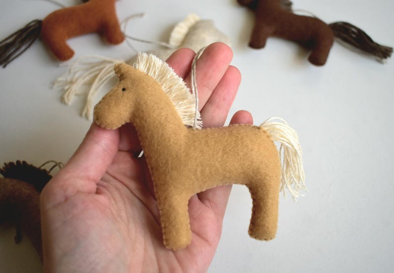 May include: Tan felt horse ornament with a cream-colored mane and tail. The horse is held in a hand, with other similar ornaments in the background. These ornaments are likely for a nursery or children's room.
