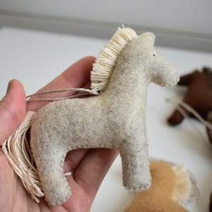 May include: A light gray felt horse ornament with a cream-colored mane and tail. The horse is held in a hand, with other felt horse ornaments in the background. The ornaments are in shades of brown and tan.
