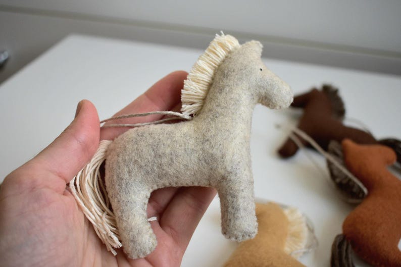May include: A light gray felt horse ornament with a cream-colored mane and tail. The horse is held in a hand, with other felt horse ornaments in the background. The ornaments are in shades of brown and tan.
