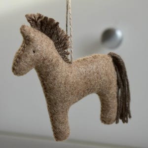 May include: A handmade, brown felt horse ornament with a dark brown mane and tail. The horse is suspended by a natural-colored string. This decorative item is suitable for nurseries, children's rooms, or as a rustic holiday decoration.