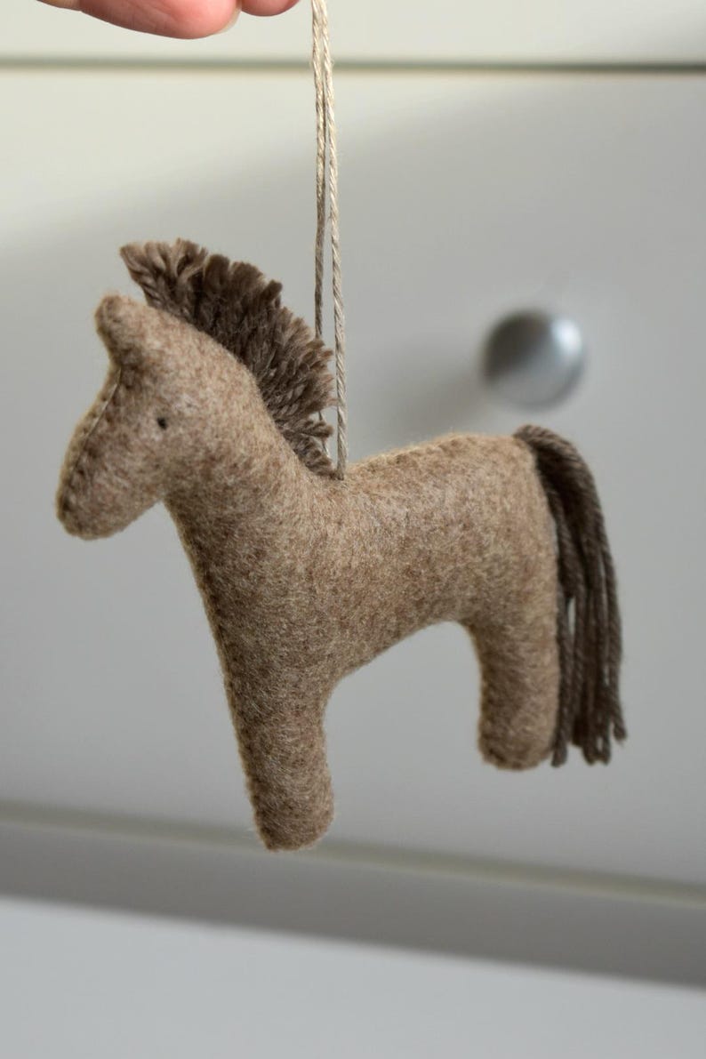 May include: A handmade, brown felt horse ornament with a dark brown mane and tail. The horse is suspended by a natural-colored string. This decorative item is suitable for nurseries, children's rooms, or as a rustic holiday decoration.