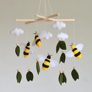 May include: A handmade baby mobile featuring yellow and black striped bees with white wings, white clouds, and green leaves. The mobile is suspended from a wooden frame with a natural finish. A charming nursery decoration.