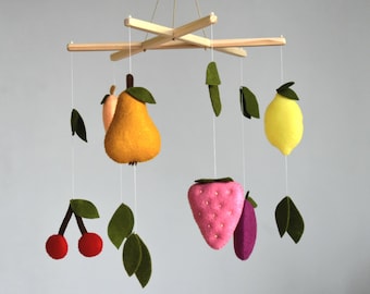 Puede incluir: Un móvil de madera con decoraciones de fruta de fieltro. El móvil presenta una pera, un limón, una fresa, una cereza y una ciruela. La fruta está suspendida de un marco de madera con hojas de fieltro verde.