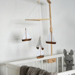 Puede incluir: Un m&oacute;vil de beb&eacute; de madera con un dise&ntilde;o de velero blanco y marr&oacute;n cuelga sobre una cuna blanca. El m&oacute;vil est&aacute; suspendido de un brazo de madera sujeto a la cuna. Un conejito de crochet gris se sienta en la cuna.