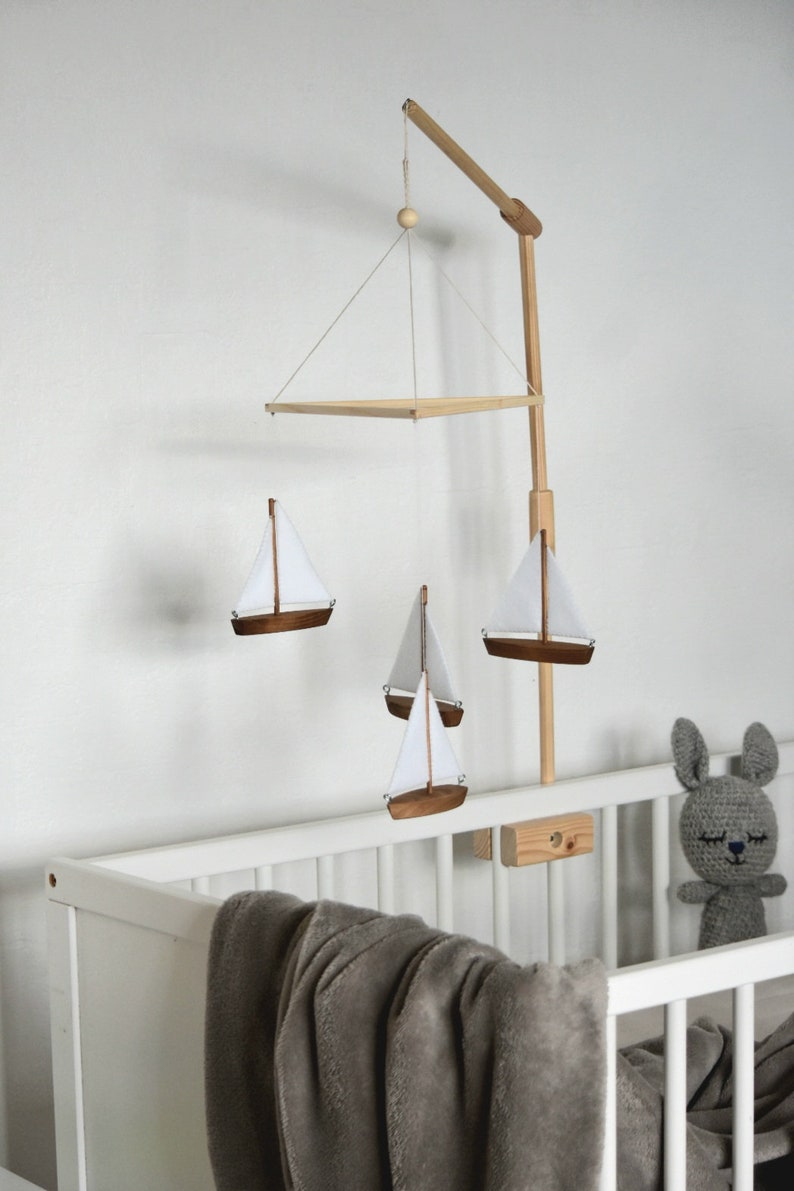 Puede incluir: Un m&oacute;vil de beb&eacute; de madera con un dise&ntilde;o de velero blanco y marr&oacute;n cuelga sobre una cuna blanca. El m&oacute;vil est&aacute; suspendido de un brazo de madera sujeto a la cuna. Un conejito de crochet gris se sienta en la cuna.