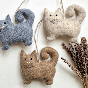 Pode incluir: Três enfeites de gato de feltro feitos à mão em cinza, bege e marrom, cada um com uma cauda enrolada e traços faciais bordados. Os gatos são suspensos por barbante e dispostos com raminhos de lavanda seca.