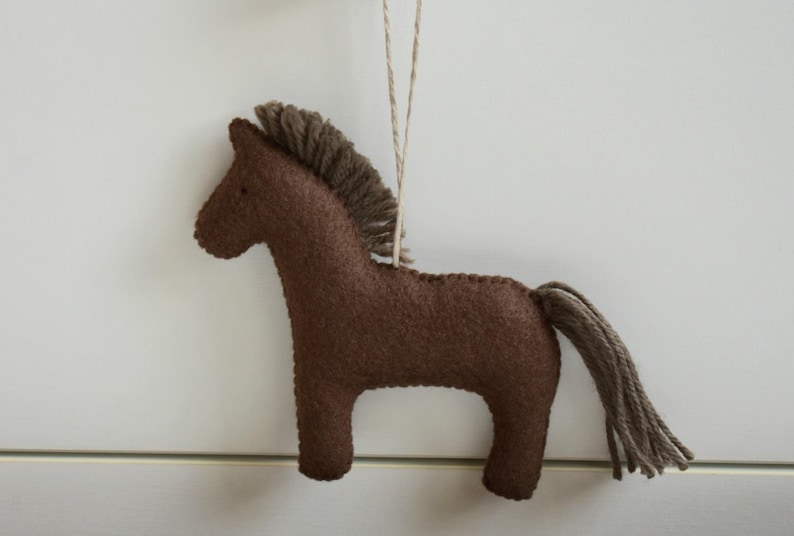 May include: A brown felt horse ornament with a brown mane and tail, hanging from a string. The horse is a simple, stylized design, perfect for nursery decor or a rustic touch to any room.