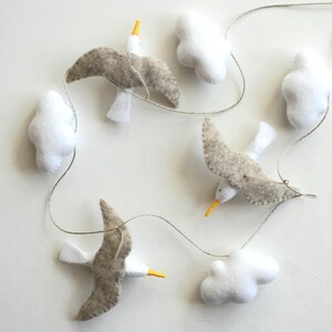 May include: A felt garland featuring three gray and white seagulls with yellow beaks and four white cloud shapes. The birds and clouds are strung on a thin, neutral-colored cord, creating a whimsical, decorative piece.