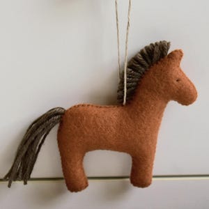 May include: A handmade felt horse ornament in shades of brown. The horse has a dark brown mane and tail, and is suspended by a thin string. The ornament is a simple, rustic design, perfect for adding a touch of charm to any space.