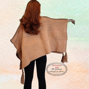 May include: A tan crochet poncho with tassels. The poncho is made of a light brown yarn and has a textured, open weave design. The tassels are a darker shade of brown. The image includes text that says "Retirement SALE All patterns reduced to clear!"