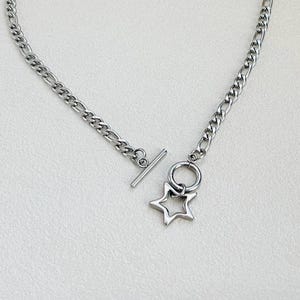 May include: A silver-colored necklace with a star pendant. The necklace features a chain with a toggle clasp closure. The star pendant is a hollow, five-pointed star shape. The necklace is displayed on a light-colored surface.
