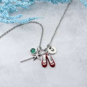 May include: Silver-toned necklace with charms including a green gem, a star, a wand, and two red shoes. A small circular charm with the letter "W" is also included. The necklace is displayed on a light gray surface.
