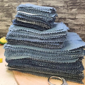 Puede incluir: Montones de cuadrados de tela vaquera azul con bordes deshilachados, listos para manualidades. Los cuadrados están cuidadosamente dispuestos sobre una tabla de cortar de madera, con un par de tijeras plateadas con mangos decorativos en primer plano. La mezclilla tiene un aspecto clásico y descolorido.