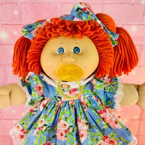 May include: A vintage doll with orange yarn hair, blue eyes, and a pacifier. The doll wears a blue floral dress with white lace trim, a matching bow, white socks, and white shoes with blue stripes. The doll is standing on a pink surface.