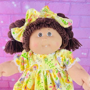 May include: A Cabbage Patch Kid doll wearing a yellow dress with a butterfly print and a yellow headband. The doll has brown yarn hair and brown eyes.