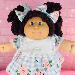 Puede incluir: Muñeca de estilo vintage con cabello rizado castaño oscuro y ojos marrones. La muñeca viste un vestido de cuadros azul y blanco con detalles florales rosas, un babero blanco y zapatos a juego. Dos lazos azules adornan el cabello de la muñeca.