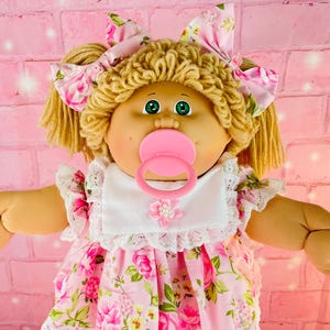 May include: A vintage doll with blonde curly hair, green eyes, and a pacifier. The doll is dressed in a pink floral dress, matching bow, and pink shoes. A white bib with lace trim and a pink flower completes the outfit.