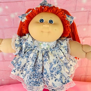 以下が含まれることがあります： 丸い顔、青い目、赤い毛糸の髪のヴィンテージ人形。 青と白の花柄のドレス、レースのトリム、白い靴下、青いストライプの白い靴を着用しています。 人形の腕と脚は柔らかく、ぬいぐるみです。