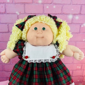 May include: A Cabbage Patch Kid doll with blonde curly hair wearing a red and green tartan dress with a white lace trim and white shoes.